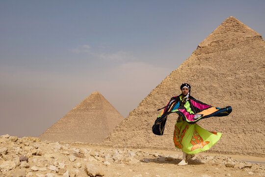 Whirling performer caught mid spin with a dramatic billowing multicolor patchwork cape against the iconic Giza Pyramids on the desert plateau in bold