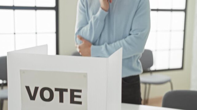 Man wearing blue sweater with hand on chin looking at vote sign and ballot booth in building; civic duty contemplation.
