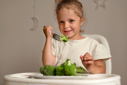 A young child sits at a table holding a fork with a piece of broccoli. She smiles while looking at the camera. The room has a simple design