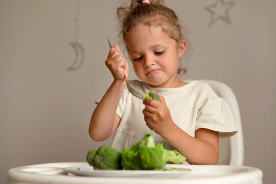 A young child sits at a table with a fork in hand. She is focused on eating broccoli. The room has soft decorations and a light feel