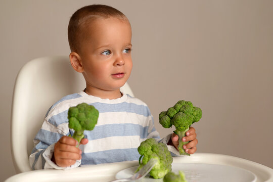 A child sits in a high chair with a focused expression, holding a piece of broccoli in each hand. The setting is bright and uncluttered