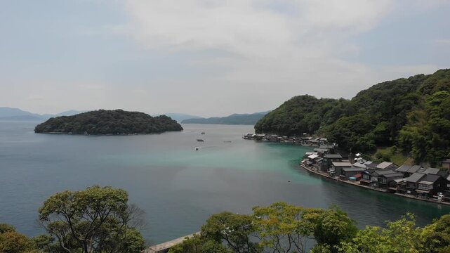 Aerial drone view descending slowly behind the trees, obscuring the serene Ine Bay and traditional Funaya boat houses in Kyoto, Japan