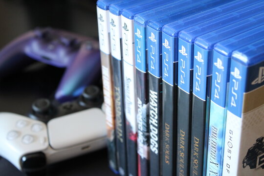 PlayStation 4 and PlayStation 5 physical game cases displayed with game controllers. Video games, gaming industry, console market, physical games, consumer electronics, entertainment business.