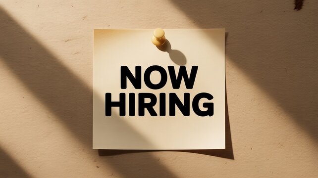 Square paper note with bold black text now hiring pinned to a textured beige wall