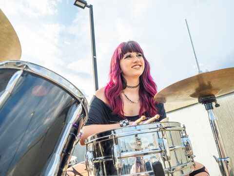 A young female drummer with vibrant purple hair poses confidently behind a drum kit while holding drumsticks outdoors, showcasing creativity, musical talent, and alternative style.