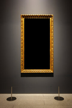 A vertical antique gilded frame with ornate details hangs on a dark museum wall above a floor with stanchions. The central black area is blank for a mockup display.