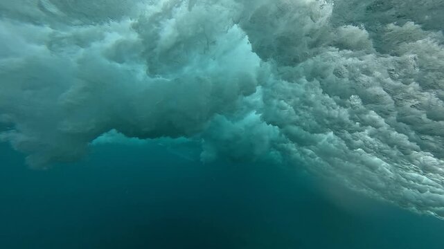 UNDERWATER: Surfer making a duck dive beneath a breaking wave at a remote surf spot in Indonesia. Foamy whitewater rolls in crystal clear blue tropical water. Power and motion of the Indian Ocean.