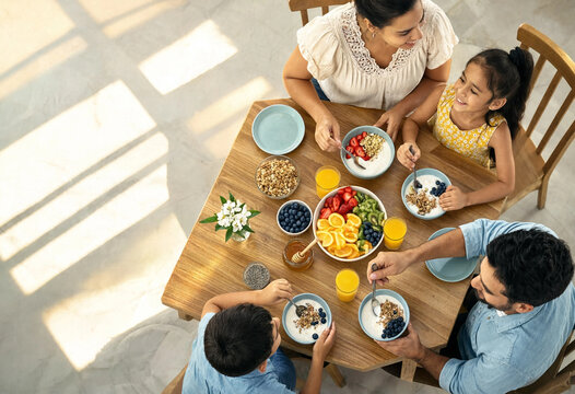 Family shares a healthy breakfast around a wooden table with bowls of fruit, yogurt, and juice. Parents and children interact warmly in bright natural light.
