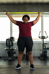 Fototapeta premium Plus size African American woman lifting green resistance band overhead in modern gym, fitness lifestyle photo for shoulder mobility, healthy workout routine, body confidence and wellness motivation.