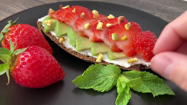 A woman's hand places a mint leaf next to a vegetarian sandwich, homemade food. Close-up of a piece of bread with avocado and strawberries. A healthy summer snack
