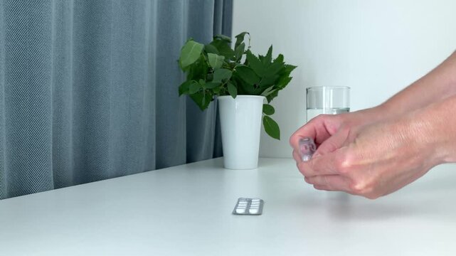 A woman takes a blister from a white table and takes out a headache pill from it. A glass of water on the background, the need to respond to symptoms of malaise in a timely manner