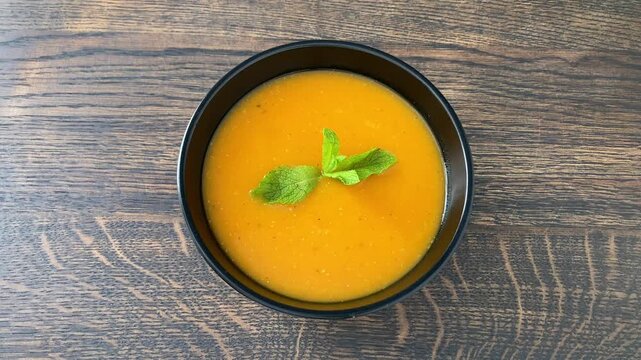 Top view of bowl of red lentil and carrot cream soup. Woman's hand puts mint leaf in black bowl with vegetarian soup, wooden background. A dish with vegetable protein, fiber, vitamins and antioxidants