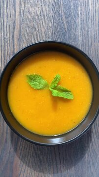 Vertical video. A woman decorates a cream soup of red lentils and carrots with a mint leaf. Top view of a black bowl of vegetarian soup on a wooden table. Healthy and balanced meal, healthy diet