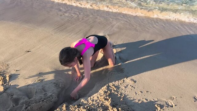 Little girl digging in wet beach sand near gentle sea waves during a sunny summer vacation. Happy childhood, outdoor play, family travel, and carefree moments by the ocean.
