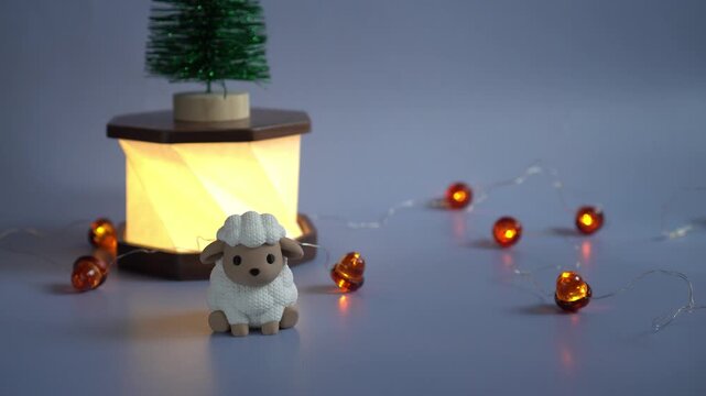 Vintage 2027 symbol white sheep figurine stands against the background of a glowing paper and wood lamp with a miniature Christmas tree on a background of flashing acorn-shaped garlands on a blue back