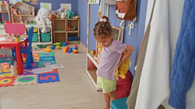 Little girl adjusts yellow bee backpack strap, barefoot in colorful classroom building; playful wonder.