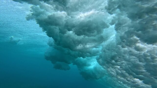 UNDERWATER: Breaking wave turns into a cloud of foam and bubbles in clear tropical water. Soft blue tones and drifting whitewater reveal raw energy and texture of a surf spot in the Mentawai Islands.