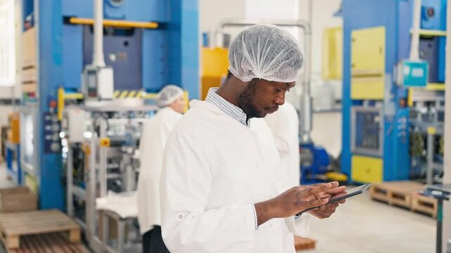 African American male supervisor in protective coat entering production info into tablet device. Focused worker monitoring manufacturing process at factory. Colleagues working in background.