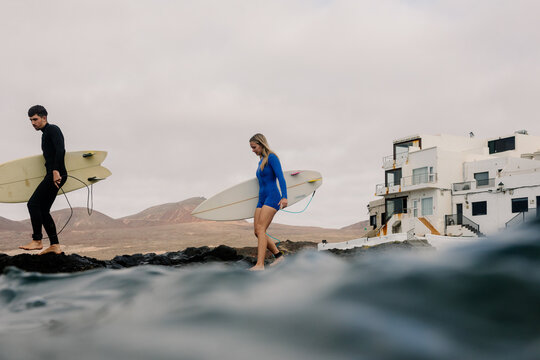 A man and a blonde woman carry surfboards along a rocky shore.