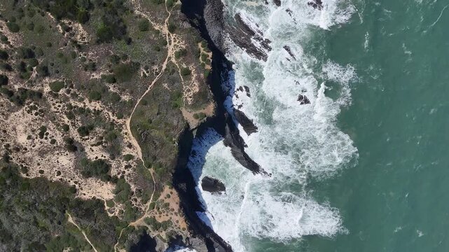 An aerial drone shot pushes in on the stunning rugged coastline near Odeceixe, Portugal. Featuring crashing waves, green vegetation, and rocky cliffs along the Atlantic Ocean.