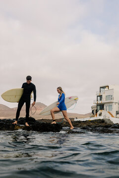 A man and a blonde woman carry surfboards across rocky terrain.