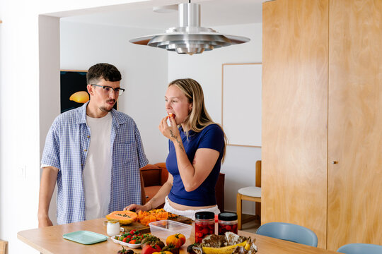 A blonde woman eats fruit while a man in a plaid shirt watches her.