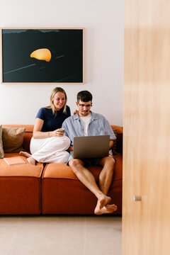 A couple relaxes on an orange couch, smiling at a laptop screen.