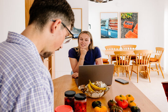 A woman with a laptop looks up at a man standing in an apartment.