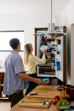 A man holds a container while a woman organizes items inside a fridge.