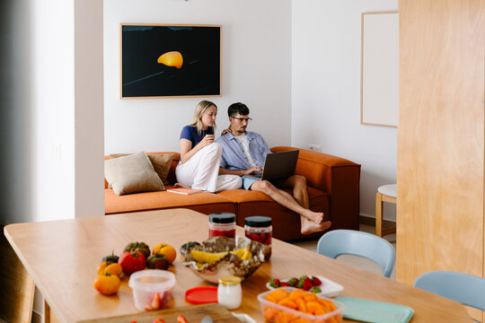 A couple relaxes on an orange sofa, watching something on a laptop.