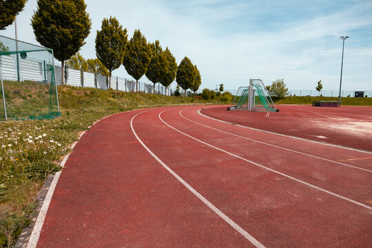Red tartan track with white lines on an outdoor sports field. Trees on the left side, blue sky with clouds. Track for training and competitions. Leisure activity, sports facility, soccer field.
