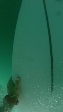 VERTICAL, FPV, SLOW MOTION: Surfer makes duck dive in front of approaching wave. Surfboard slices underwater, surrounded by bubbles and turquoise water. Rider paddles back out through breaking waves