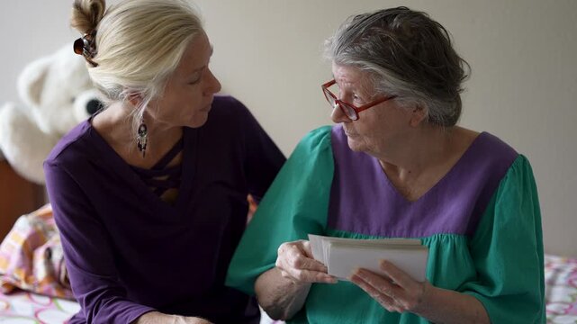 A mature daughter visits her elderly mother in a senior care facility. They share stories and look at photos.