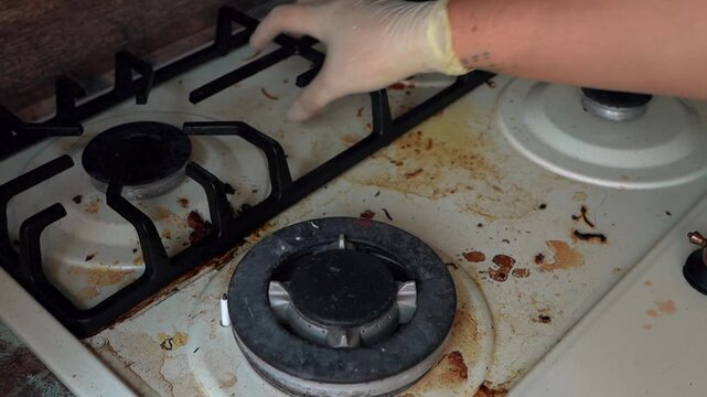 Dirty gas stove with burnt residue and food splatters, enamel burners stained with grease, gloved hand beginning cleanup, dim domestic kitchen counter visible, urgent deepclean scene