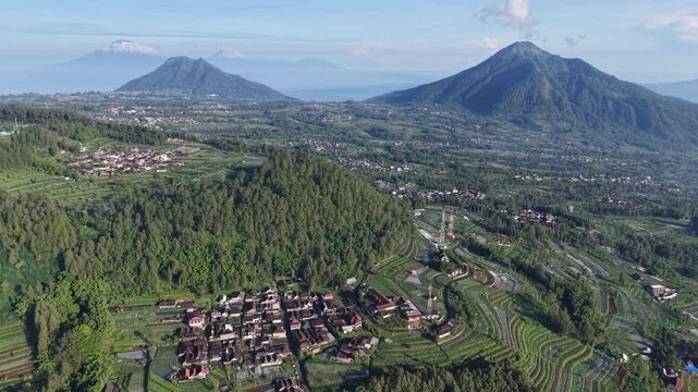 Aerial panorama of a rural Central Java village surrounded by terraced farms, lush green hills and volcanic mountains, showcasing agricultural landscape, morning light and serene, scenic countryside