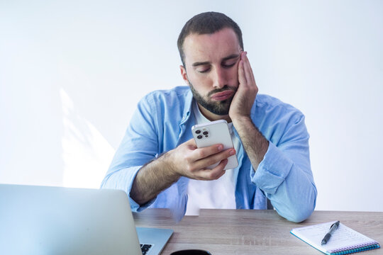 Bored man at desk looking at smartphone during remote work with laptop and notebook, feeling distracted and frustrated
