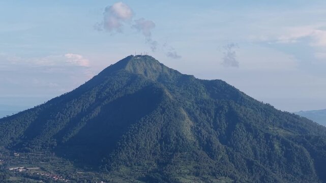 A serene forested mountain peak with small summit antennas rising above a valley, bathed in soft morning light. Lush ridges and deep shadows create a peaceful, majestic landscape scene.