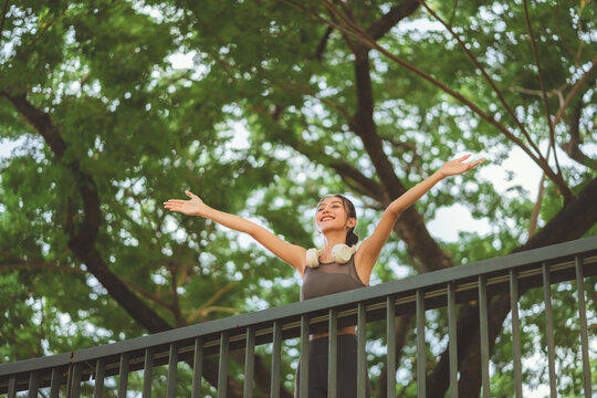 Low angle view of happy Asian woman stretching arms and breathing fresh air at green park, smiling fitness female athlete relaxing after outdoor workout for healthy wellness, vitality, active urban