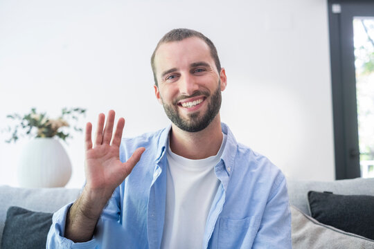 Smiling man waving while sitting on a sofa at home, greeting someone during a video call or friendly conversation