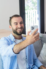 Fototapeta premium Young man smiling and taking a selfie on smartphone while relaxing on a couch at home, wearing a casual shirt and enjoying a video call or social moment