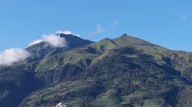 Lush green volcanic slopes and terraced hills beneath a clear blue sky with drifting clouds, capturing the serene, rugged landscape of Central Java, Indonesia ideal for travel, nature, and outdoor
