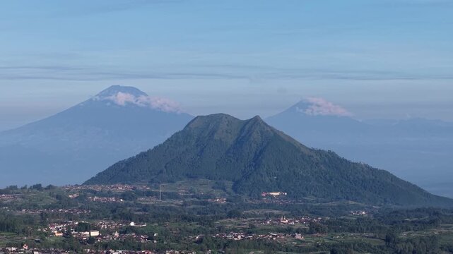 Panoramic view of Central Java volcanic peaks and verdant hills overlooking scattered rural villages under a clear blue morning sky, conveying peacefulness, natural beauty and dramatic mountainous