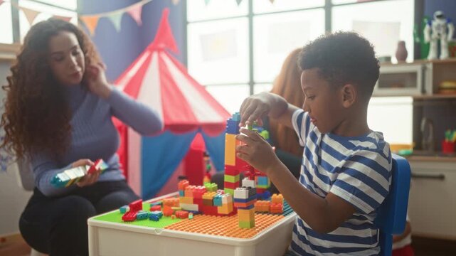 Woman and boy stacking colorful plastic blocks with hands at a play table in a school building; concentration learning.
