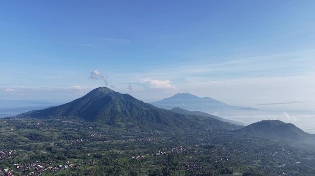 Expansive panoramic view of volcanic peaks rising above a green valley in Central Java, Indonesia, with soft morning light, clear blue sky and distant mist creating a peaceful, natural atmosphere.