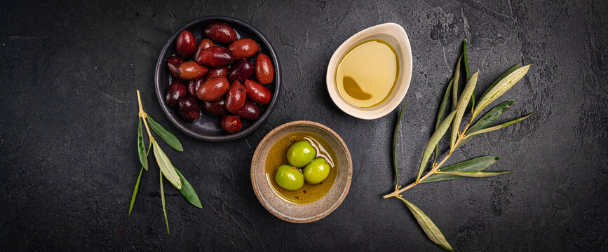 Green and black olives in a bowls and olive oil on dark background top view. Olive oil and variety of green and black olives in bowls with olive leaves all around taken from above.