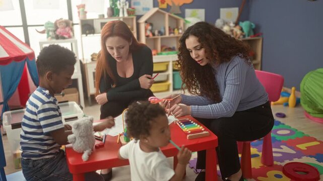 Women teaching boy and little girl at red table, handing wooden xylophone mallet in classroom; nurturing play learning.