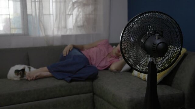 Electric fan cooling woman resting on sofa during extreme summer heat at home