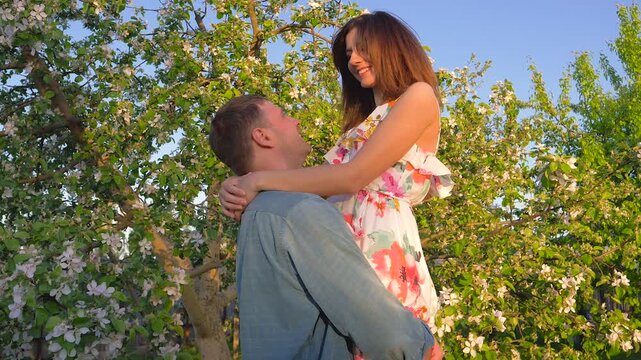 Young handsome man holding then raising and spinning his beautiful laughing girlfriend in a flourishing apple orchard. Loving couple enjoying romantic date outdoors on sunny day with clear blue sky