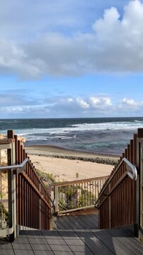 Stairs lead down to a sandy beach and ocean with waves. Clouds are in the sky above the water.