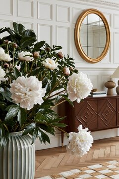 White peonies in a decorative vase with green leaves, elegantly arranged in a stylish interior featuring a round mirror and wooden sideboard